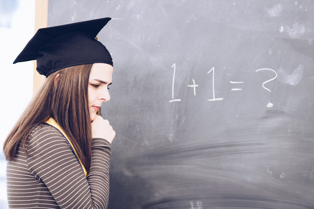 Melissa Hazlewood, a brunette girl with a graduation cap and sash on and a confused face at a chalkboard with '1+1=?' on it.